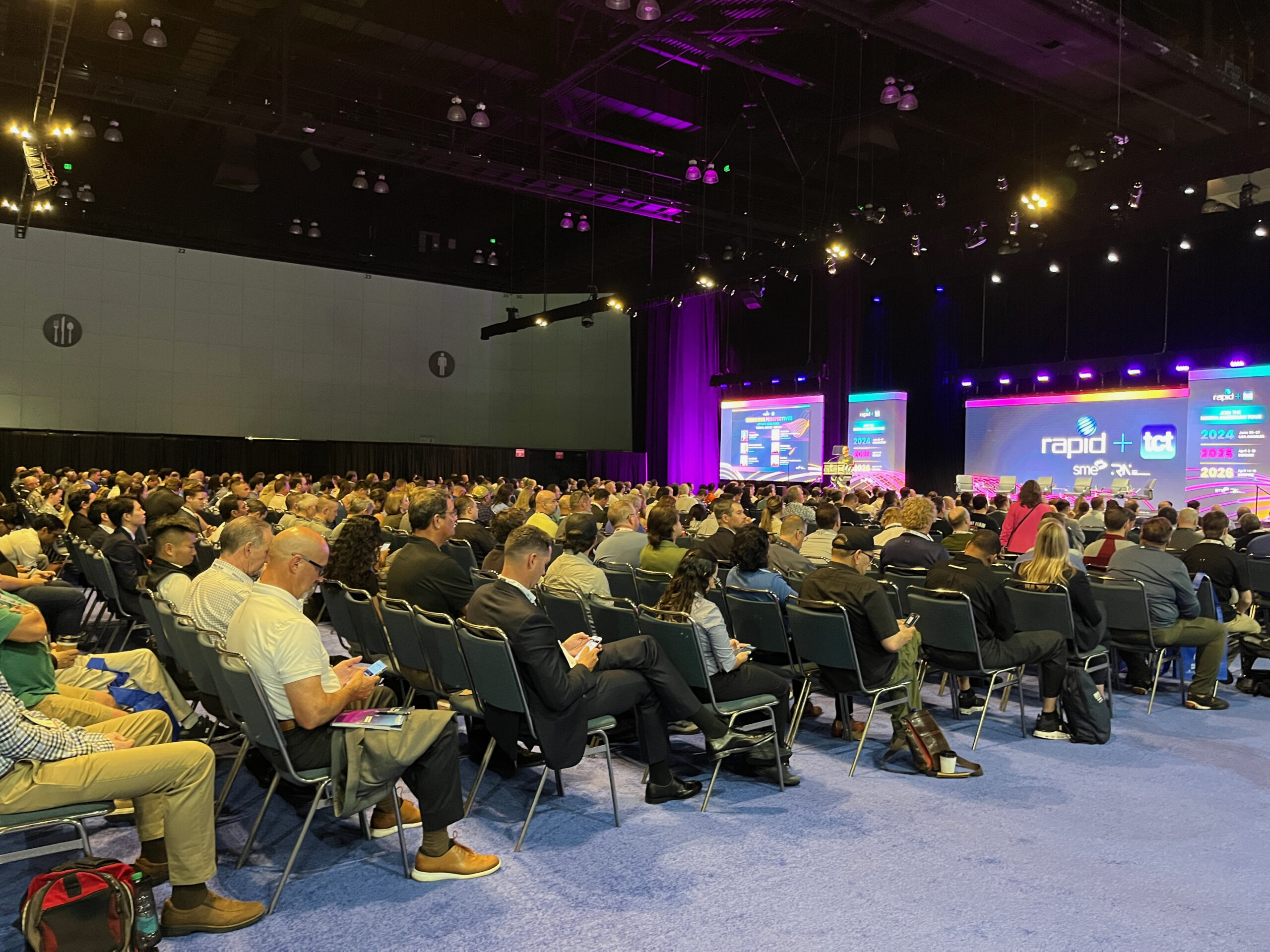 A large auditorium with thousands of chairs is occupied by many people. A stage is at the front with screens with the conference logo.