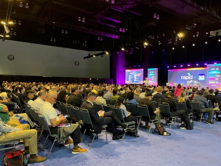 A large auditorium with thousands of chairs is occupied by many people. A stage is at the front with screens with the conference logo.