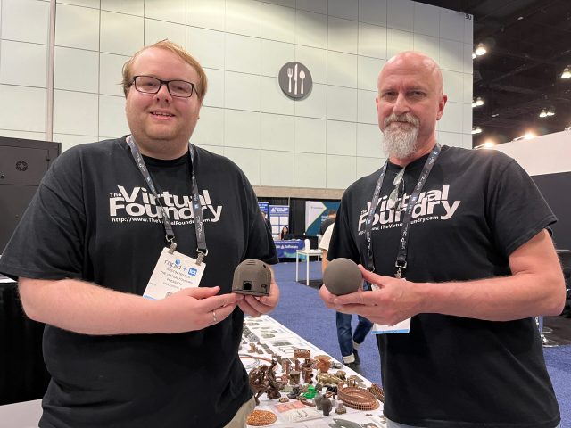 Two men in Virtual Foundry t-shirts are holding fist-sized objects that look like small domes for habitats on other planets. They are in a convention hall and their booth with many models is behind them.