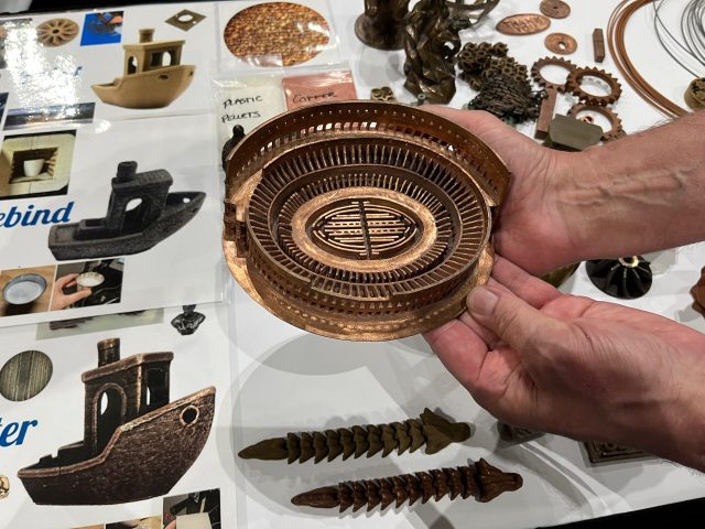 A pair of hands holds a copper model of the Roman Colosseum. Other models and photos of models are in the background.