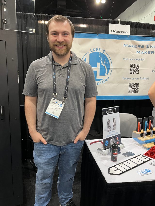 A man is smiling broadly next to a refrigerator-sized 3D printer cabinet. A sign behind him advertises Luke’s Laboratory with QR codes for his website.