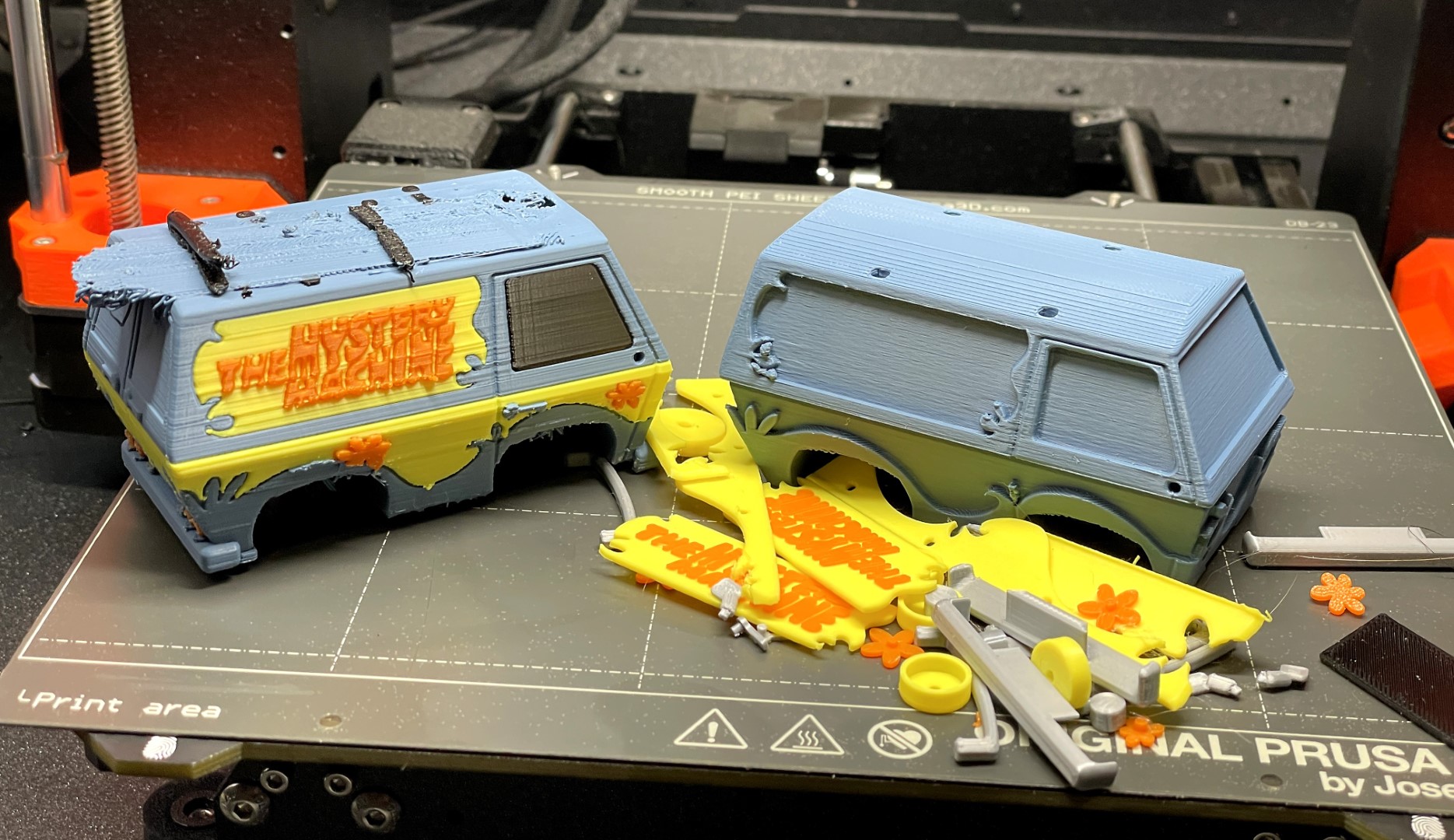 Two 3D printed van models, sitting on a printer bed. The left van looks complete, although the top is badly smeared. The right van has a pile of parts beside it waiting to be assembled.